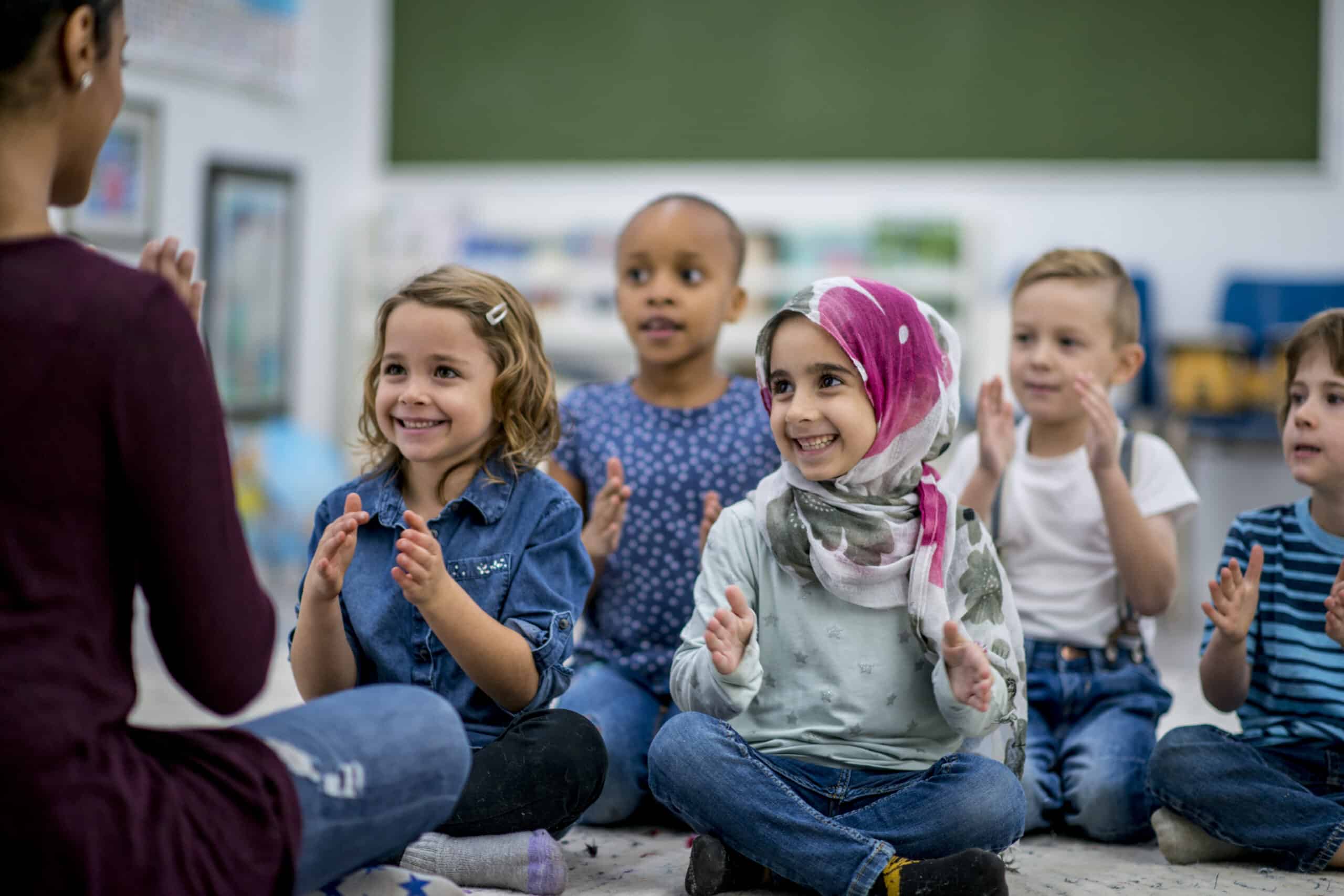 A multi-ethnic group of young school children are indoors in their classroom. They are sitting on the floor and clapping along to music with their teacher.