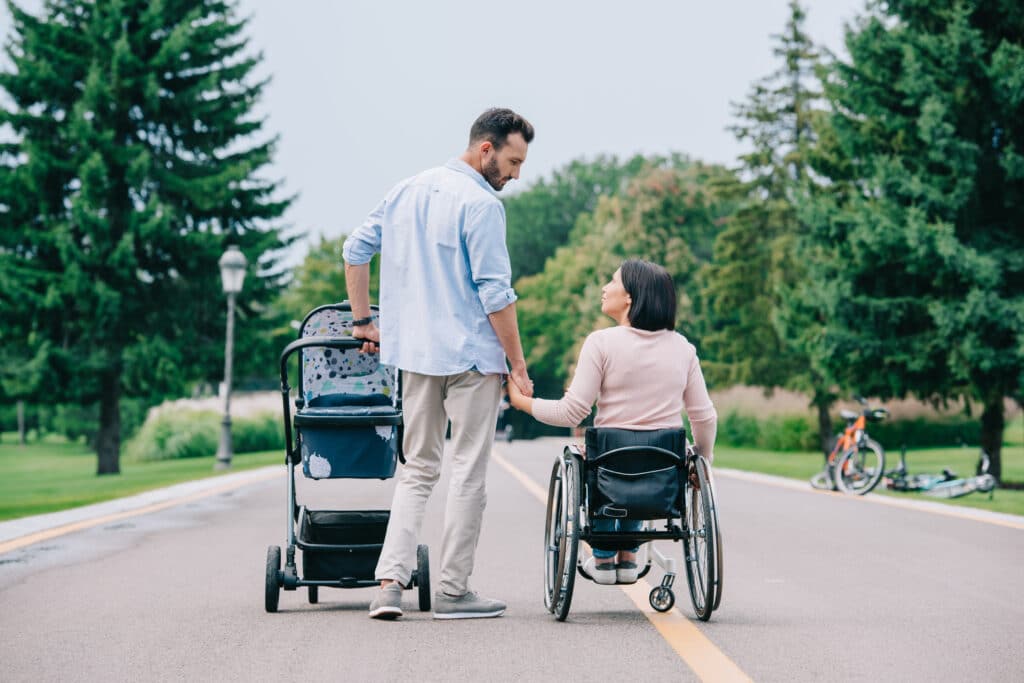 happy man holding hand of disabled wife while walking with baby carriage in park together