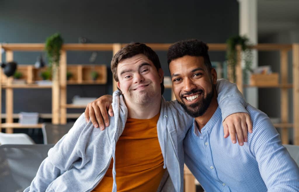 Young man with Down syndrome and his tutor with arms around each other looking at camera indoors at school