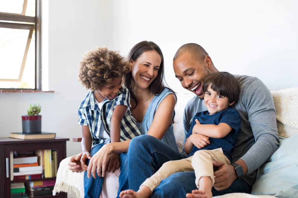 Happy multiethnic family sitting on sofa laughing together. Cheerful parents playing with their sons at home. Black father tickles his little boy while the mother and the brother smile